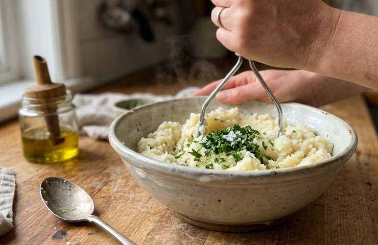 Preparazione del purè di sedano rapa fatto in casa: mani che schiacciano la radice cotta con un passaverdura in una ciotola.