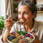 Ritratto ravvicinato di una donna sorridente in cucina mentre assaggia una bowl colorata ricca di fibre prebiotiche: si distinguono legumi neri, cereali antichi, semi di chia e mirtilli per la salute intestinale e il Fiber-maxxing.