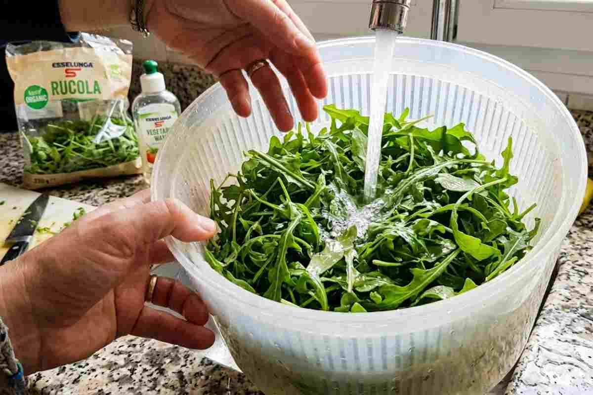 Primo piano realistico e luminoso in una cucina domestica mentre si lavano foglie fresche di rucola sotto l'acqua corrente del rubinetto, all'interno di una centrifuga per insalata. Una mano umana agita la verdura. Sullo sfondo, una busta di rucola aperta e un lavello da cucina.