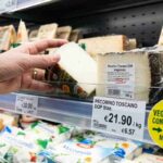 Primo piano di una mano che sceglie un formaggio Pecorino tra gli scaffali del supermercato, con un'etichetta ingrandita che evidenzia la parola 'caglio' e un bollino giallo informativo per la spesa vegetariana consapevole.