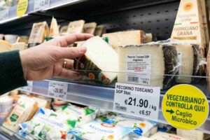 Primo piano di una mano che sceglie un formaggio Pecorino tra gli scaffali del supermercato, con un'etichetta ingrandita che evidenzia la parola 'caglio' e un bollino giallo informativo per la spesa vegetariana consapevole.