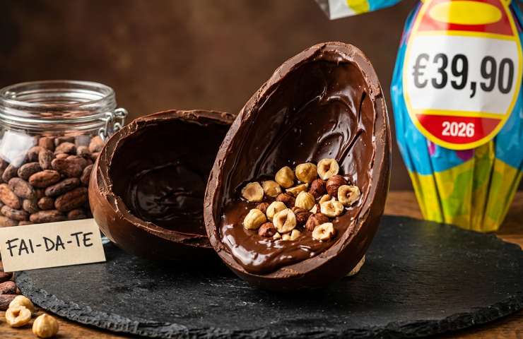 Uovo di Pasqua artigianale fai-da-te con cioccolato fondente e nocciole intere su ardesia, accanto a un barattolo di fave di cacao e un uovo industriale confezionato con cartellino del prezzo alto (39,90€, 2026). Questa immagine iperrealistica in formato 16:9 mostra il contrasto tra la preparazione domestica economica e i rincari alimentari dei prodotti da supermercato per Pasqua 2026.