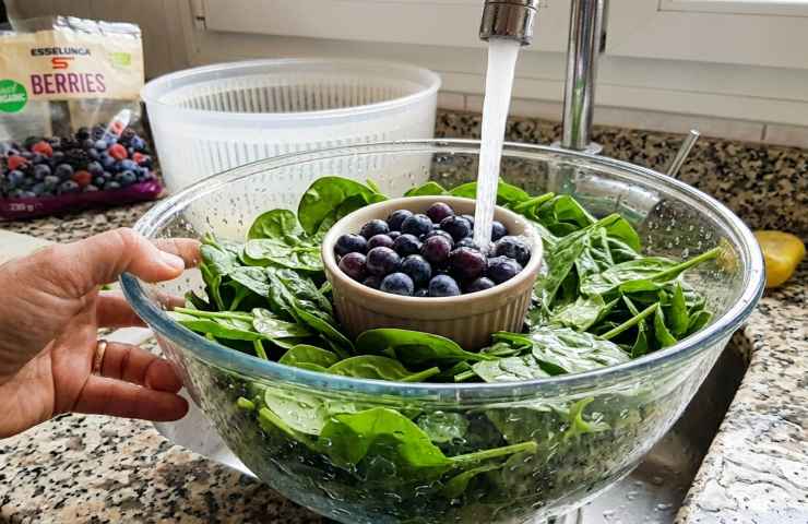 Lavaggio sotto acqua corrente di spinaci freschi e mirtilli in ciotola di vetro in cucina. Primo piano realistico con mano, fede e braccialetto.