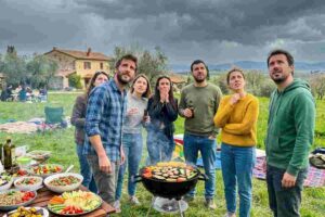 Gruppo di amici preoccupati durante una grigliata all'aperto in Italia, mentre guardano il cielo nuvoloso che minaccia pioggia. Sulla tavola, insalate miste, verdure e pane.