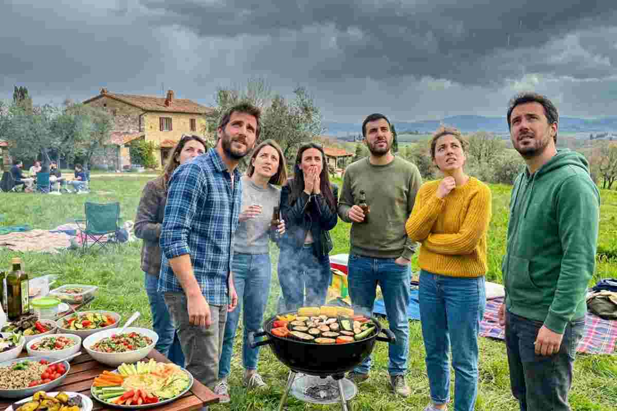 Gruppo di amici preoccupati durante una grigliata all'aperto in Italia, mentre guardano il cielo nuvoloso che minaccia pioggia. Sulla tavola, insalate miste, verdure e pane.