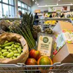 Primo piano reale di un carrello della spesa di Pasqua 2026 con fave fresche, asparagi, pecorino vegetale e colomba al pistacchio in un supermercato luminoso.
