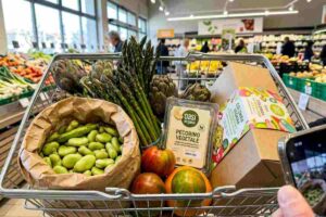 Primo piano reale di un carrello della spesa di Pasqua 2026 con fave fresche, asparagi, pecorino vegetale e colomba al pistacchio in un supermercato luminoso.