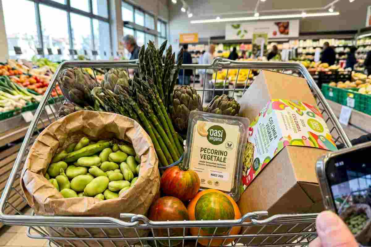 Primo piano reale di un carrello della spesa di Pasqua 2026 con fave fresche, asparagi, pecorino vegetale e colomba al pistacchio in un supermercato luminoso.