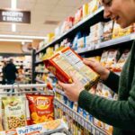 Donna che legge l'etichetta di biscotti Speculoos in un supermercato italiano con carrello pieno di prodotti della spesa vegana accidentale come taralli, patatine alla paprika e pasta sfoglia.