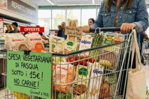 Donna spinge un carrello della spesa vegetariana di Pasqua 2026 con prodotti MDD e cartello spesa sotto i 15 euro nel reparto freschi del supermercato.