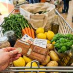 Mano con anello d'argento posa monete in portafoglio tra verdure fresche e legumi in carrello spesa.