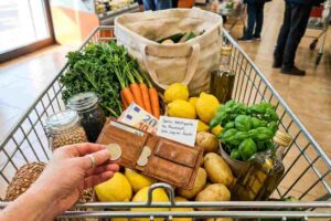 Mano con anello d'argento posa monete in portafoglio tra verdure fresche e legumi in carrello spesa.